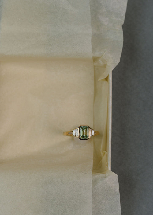 A yellow gold ring with a green diamond emerald center stone and white diamond side stones on a pale yellow background. (view 6)