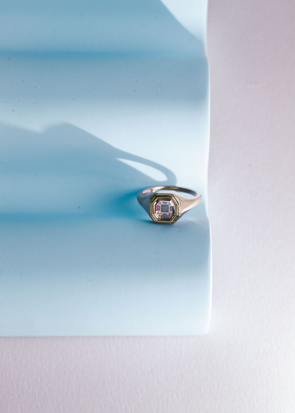 A gold signet ring with a portrait cut diamond and multi colored gemstones, resting on a light blue textured paper prop against a pale pink background. (view 4)
