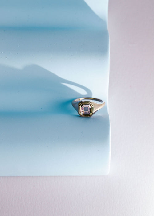 A gold signet ring with a portrait cut diamond and multi colored gemstones, resting on a light blue textured paper prop against a pale pink background. (view 4)
