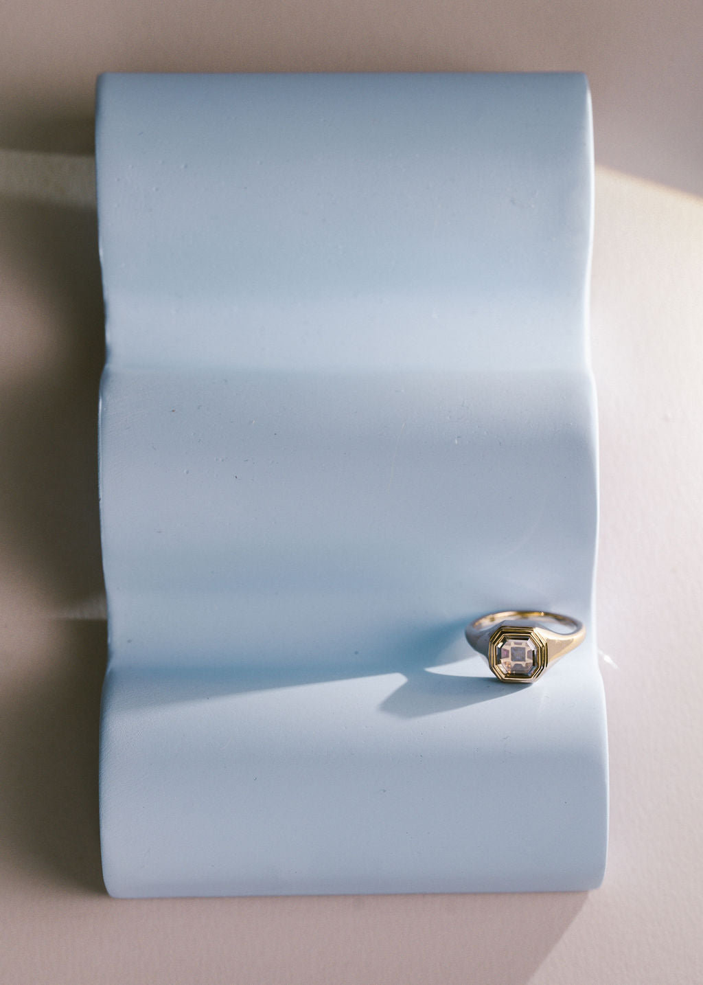 A gold ring with a square clear gemstone displayed on a light blue wavy stand against a beige background. (view 7)
