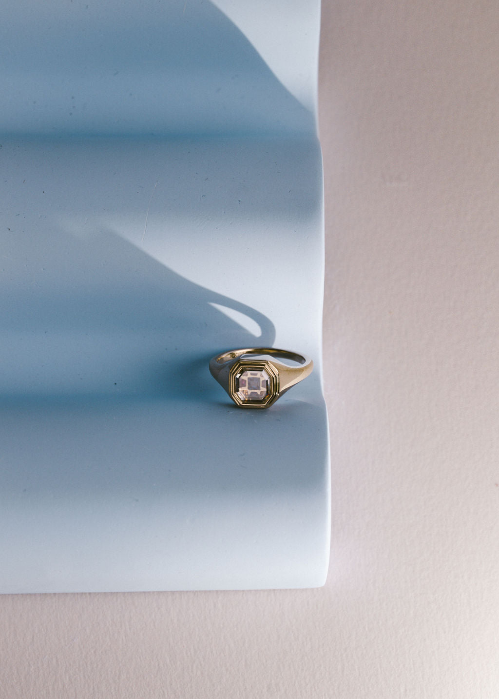 A gold signet ring with an portrait cut clear gemstone, displayed on a light blue textured paper prop against a neutral beige background. (view 10)
