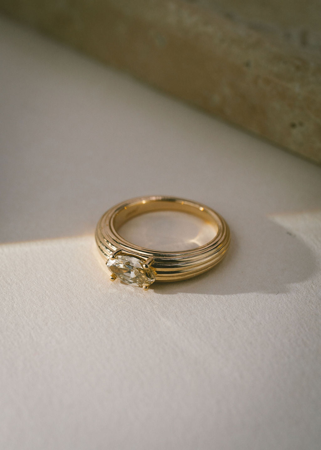 A textured gold ring with an oval champagne gemstone, resting on a textured white surface with soft shadows. (view 2)