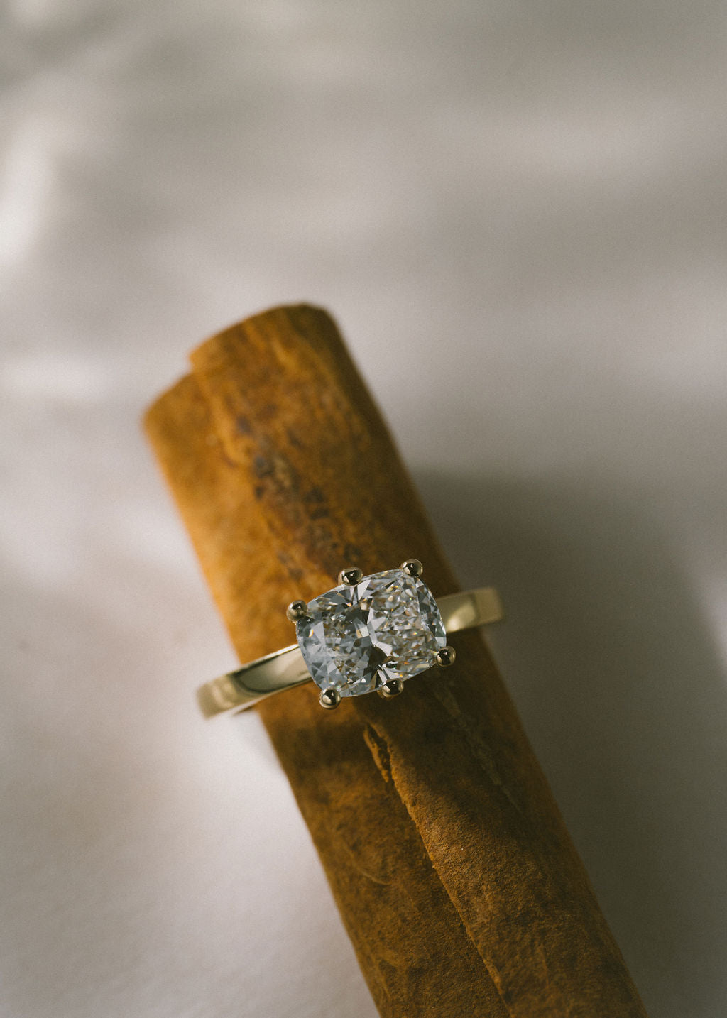 A gold ring with a cushion-cut diamond solitaire setting, displayed on a textured wooden dowel against a soft, light gray fabric background.
