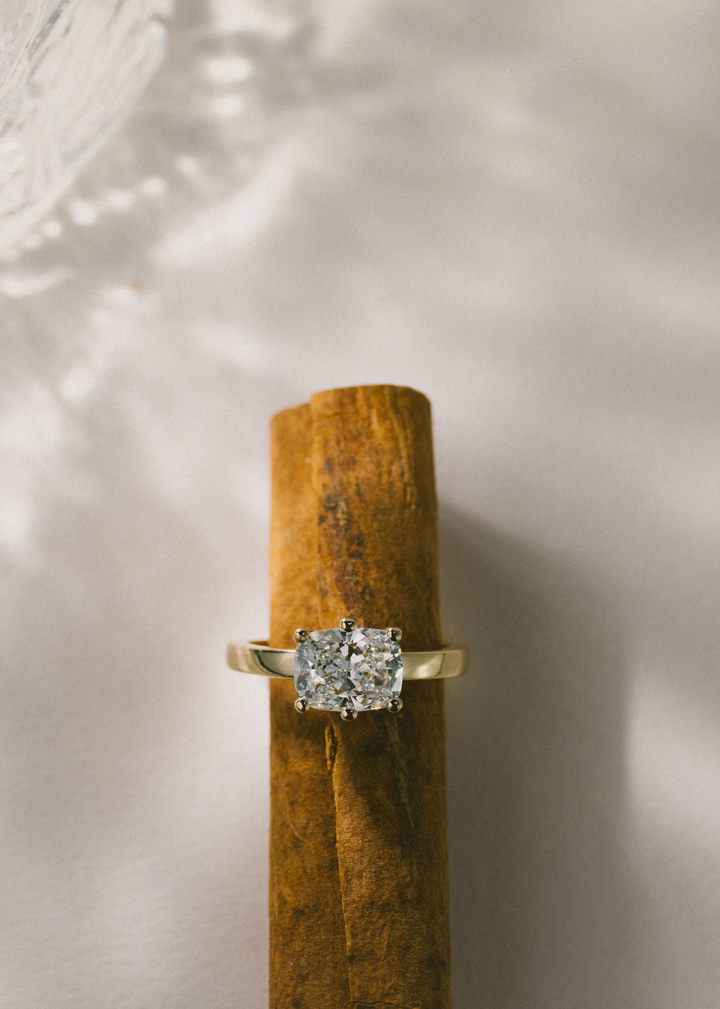 A yellow gold ring with a cushion diamond, displayed on a textured brown cinnamon stick against a soft, light gray background with leaf shadows. (view 9)