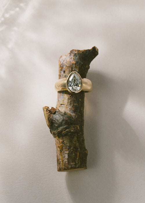 A gold ring with a pear-cut clear gemstone displayed on a small piece of natural wood against a soft white background. (view 3)