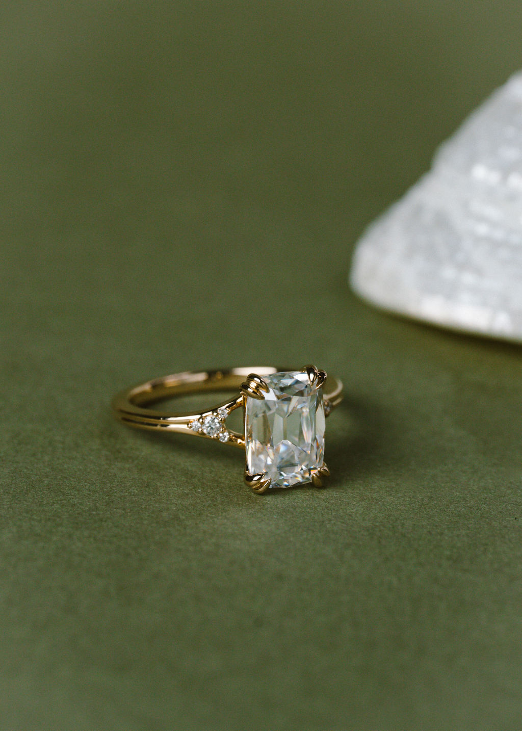 A gold ring with an cushion clear center stone and small side stones, photographed on a dark olive green surface with a blurred white crystal in the background.