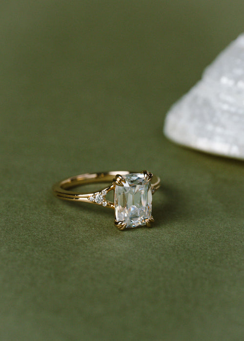 A gold ring with an cushion clear center stone and small side stones, photographed on a dark olive green surface with a blurred white crystal in the background.