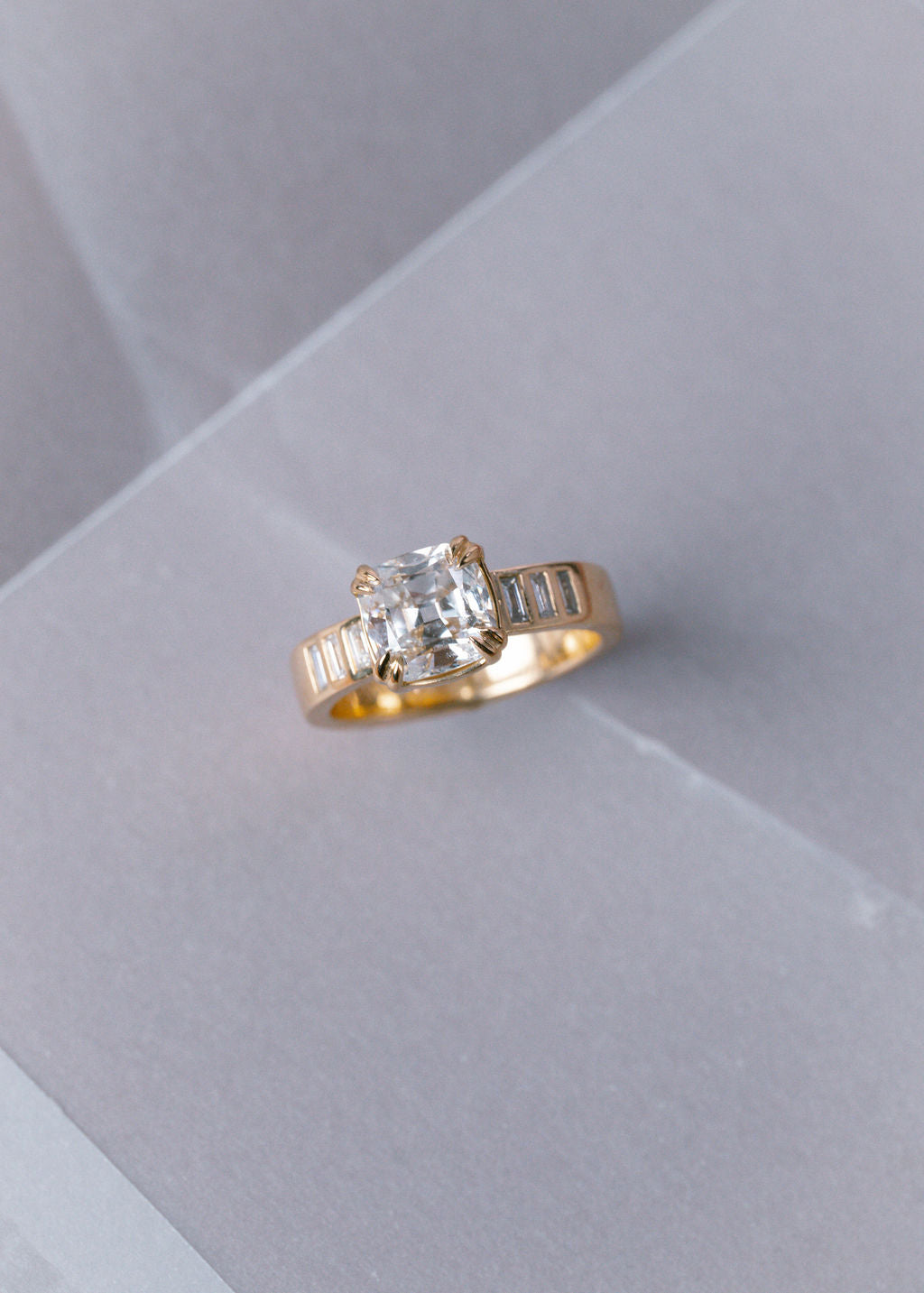 A yellow gold ring featuring a cushion-cut clear center stone and baguette side stones, displayed on a light gray textured surface. (view 2)