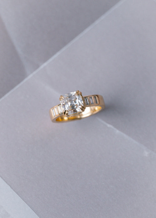 A yellow gold ring featuring a cushion-cut clear center stone and baguette side stones, displayed on a light gray textured surface.