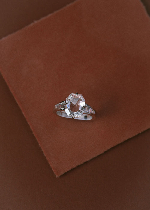 A silver ring with a large clear diamond and smaller side stones, displayed on a textured brown suede-like square against a matching brown background. (view 2)
