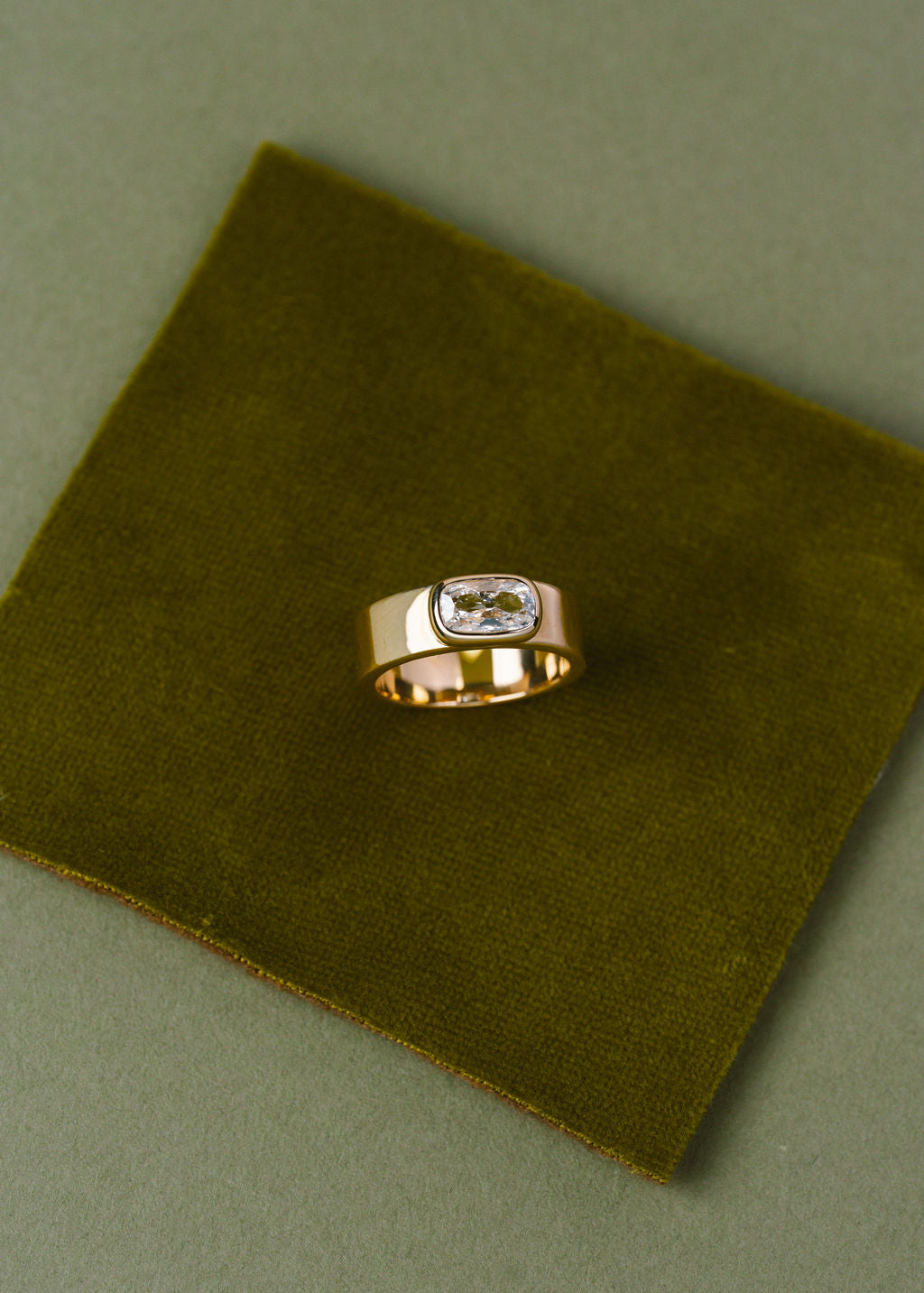 A gold ring with a cushion-cut clear gemstone, displayed on a square olive green velvet fabric card against a matching olive green background.
