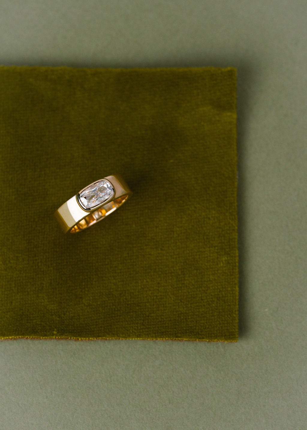 A gold ring with a bezel-set clear cushion gemstone, displayed on a square olive green velvet fabric pad against a matching olive green background. (view 4)