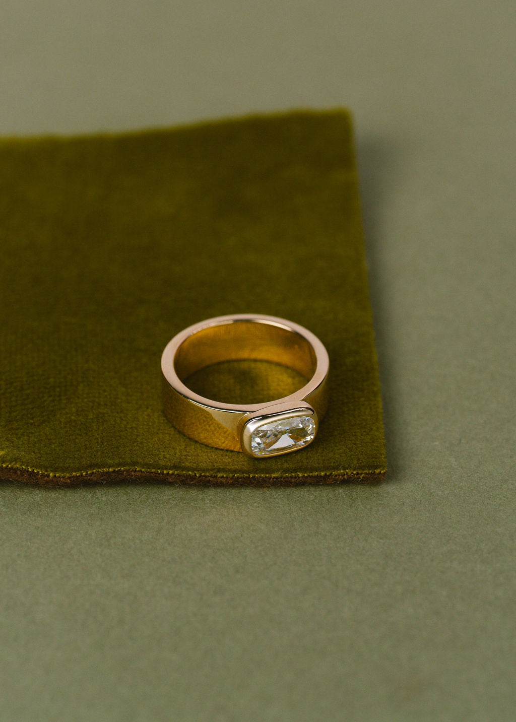 A yellow gold ring with a clear cushion gemstone, resting on a folded olive green velvet fabric against a solid olive green background. (view 7)