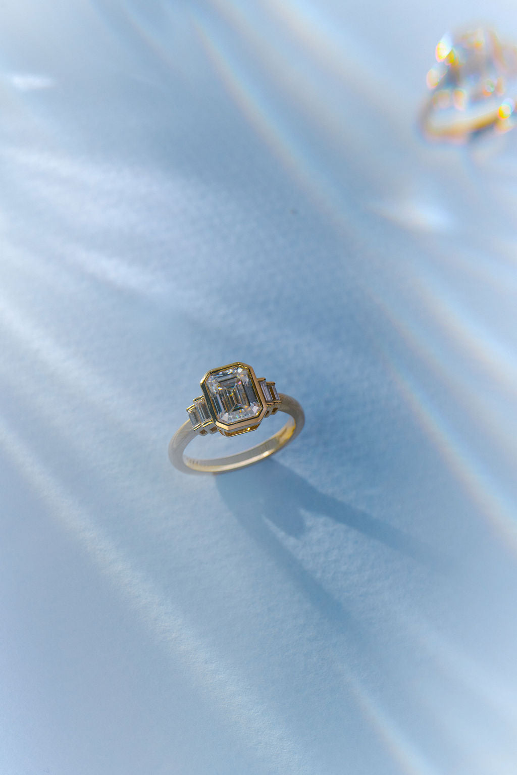 A gold ring with a geometric emerald-cut clear center stone, resting on a light blue satin fabric background.