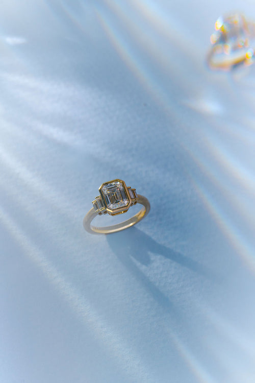 A gold ring with a geometric emerald-cut clear center stone, resting on a light blue satin fabric background.