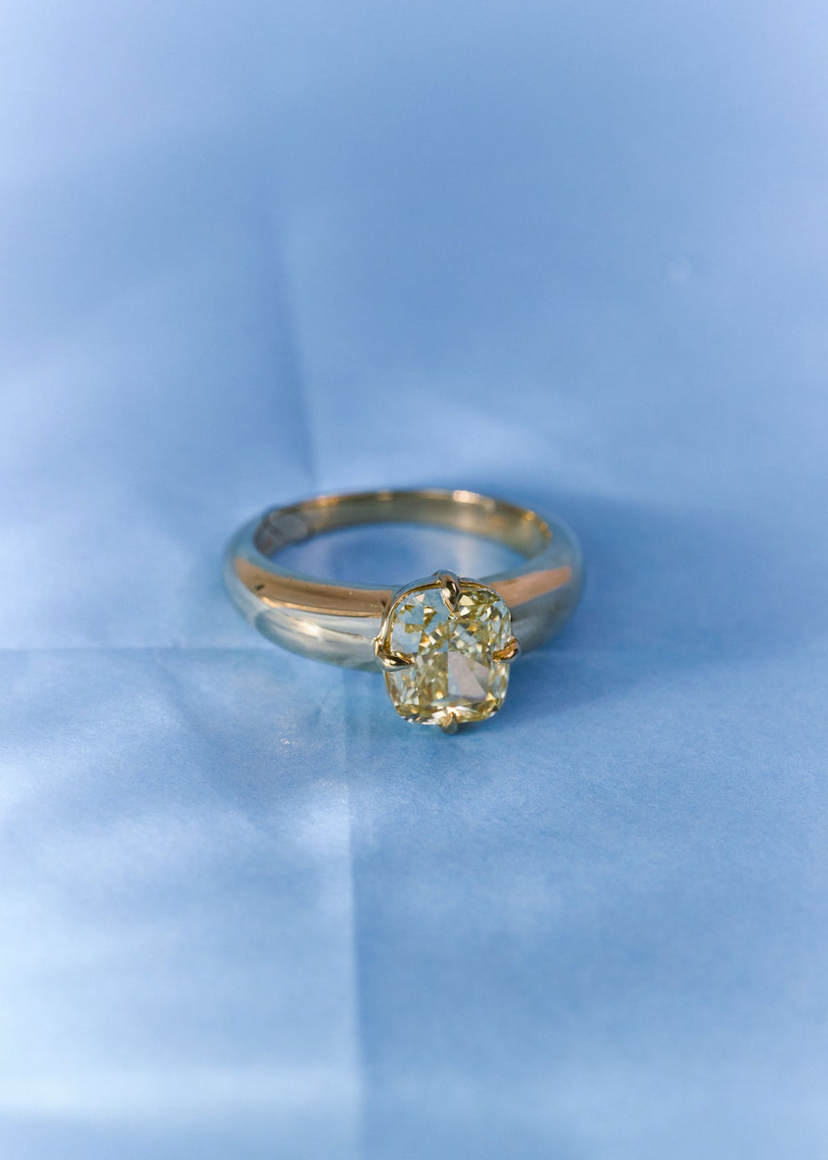 A  yellow gold solitaire ring with a yellow diamond, resting on a textured light blue surface.