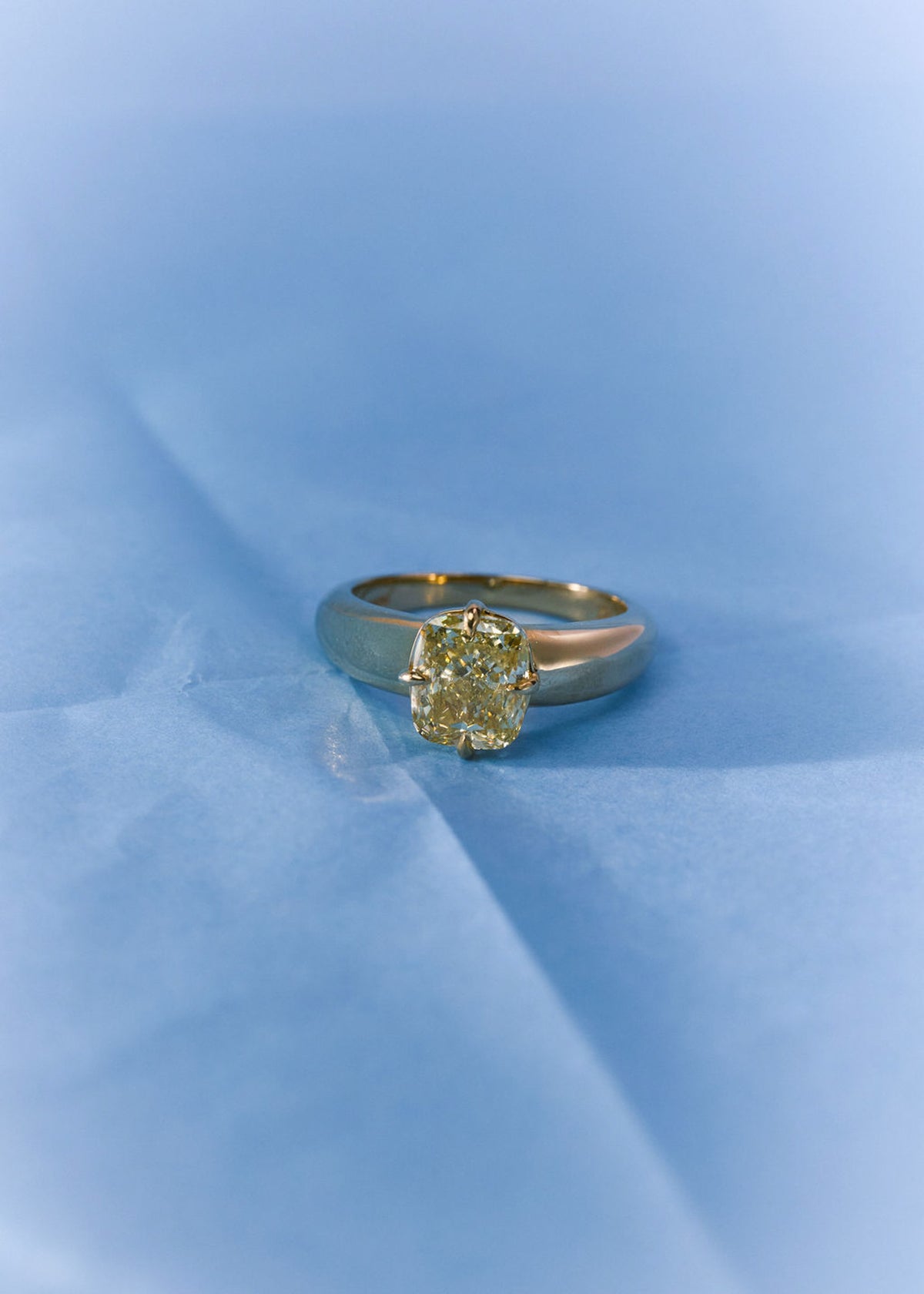 A yellow gold ring with a yellow gemstone, resting on a soft, light blue textured surface. (view 9)