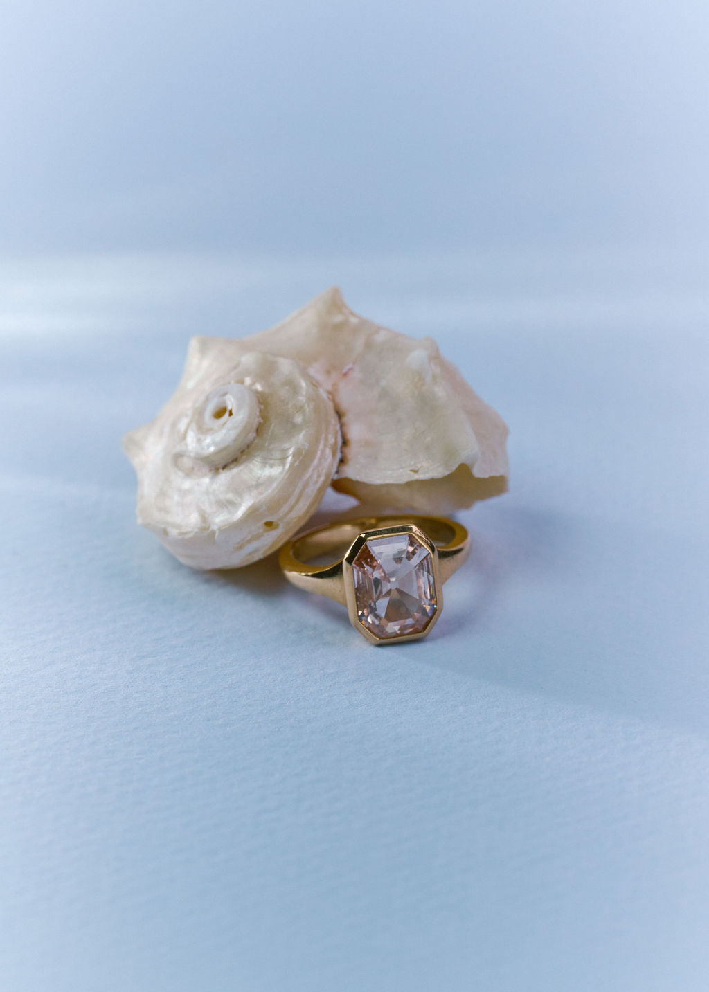 A gold ring with an octagonal  champagne gemstone set in a gold bezel, displayed next to a large white seashell on a light blue background. (view 7)
