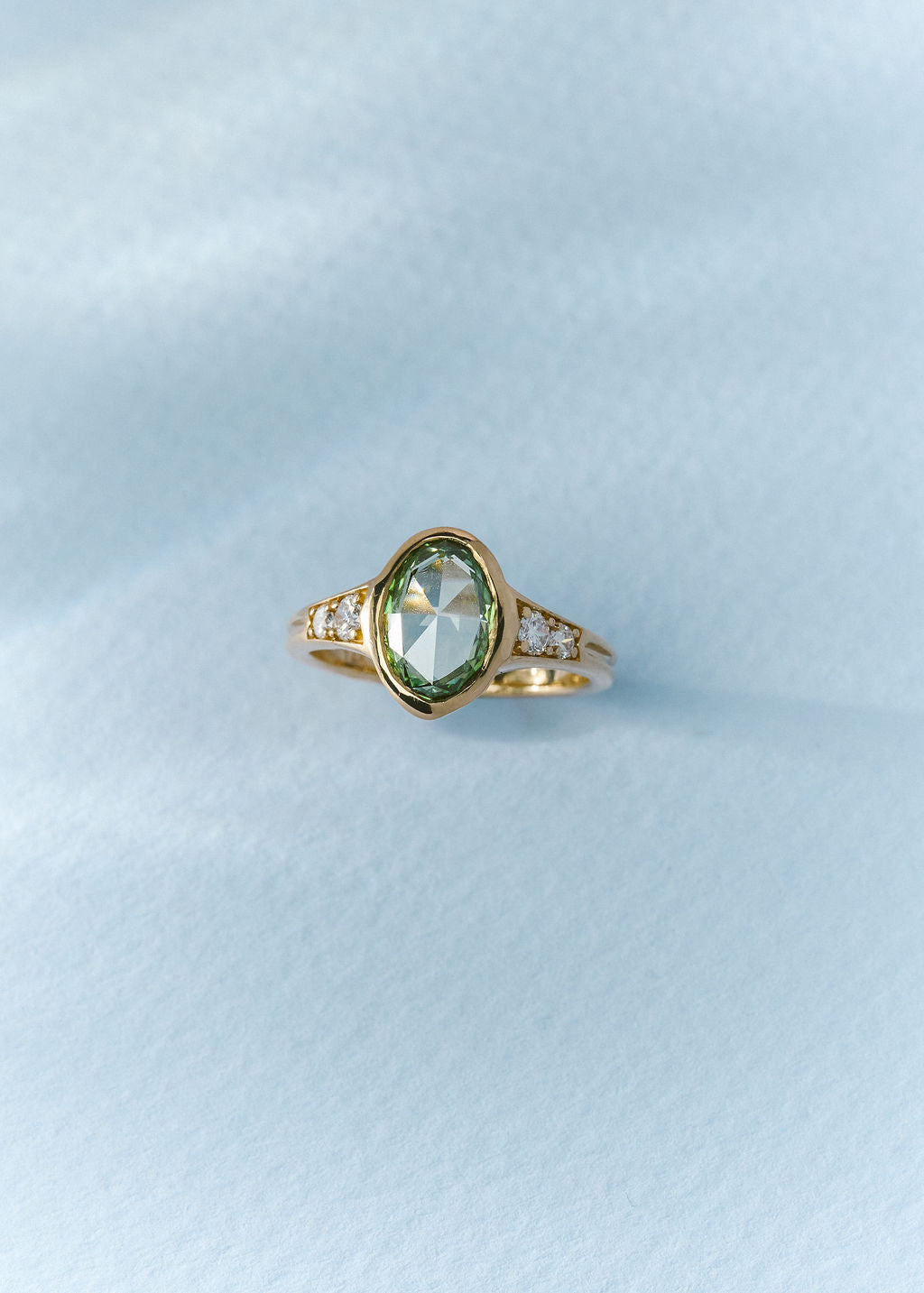 A gold ring featuring a rose cut oval green gemstone in a bezel setting, with small clear accent stones on either side of the band, resting on a textured light blue surface.