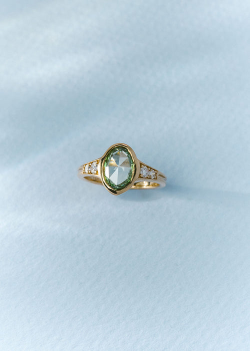 A gold ring featuring a rose cut oval green gemstone in a bezel setting, with small clear accent stones on either side of the band, resting on a textured light blue surface.