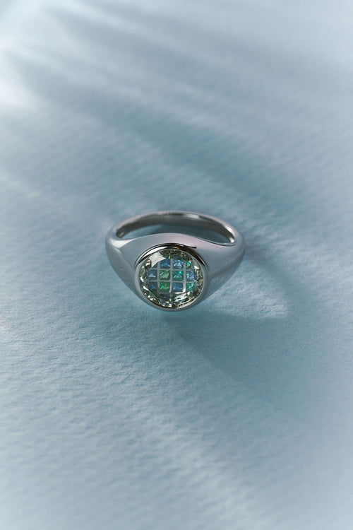 A silver signet ring with a round face featuring a green and blue checkered pattern, resting on a textured light blue surface.