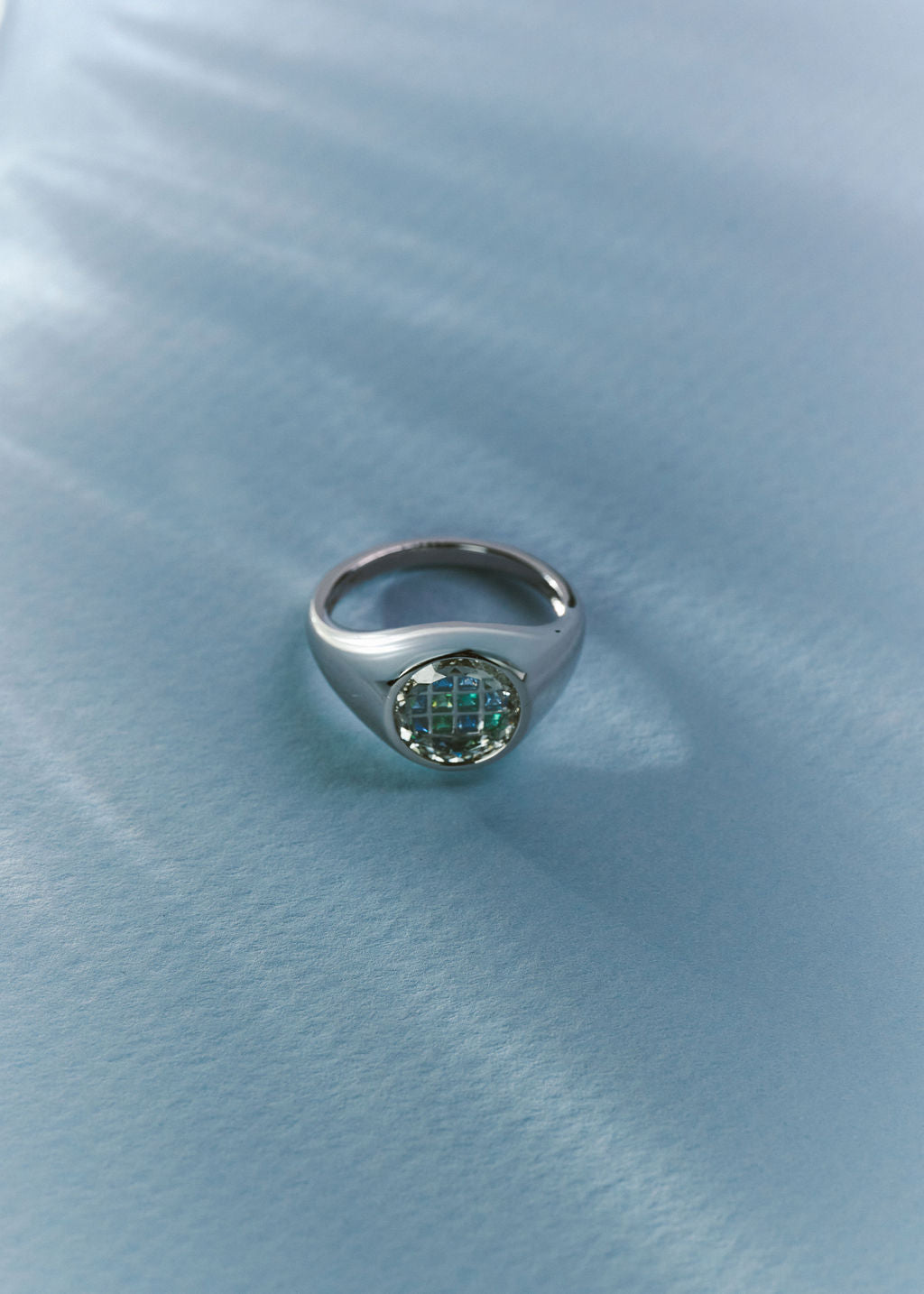 A silver signet ring with a round, textured green gemstone, resting on a textured light blue fabric background. (view 4)