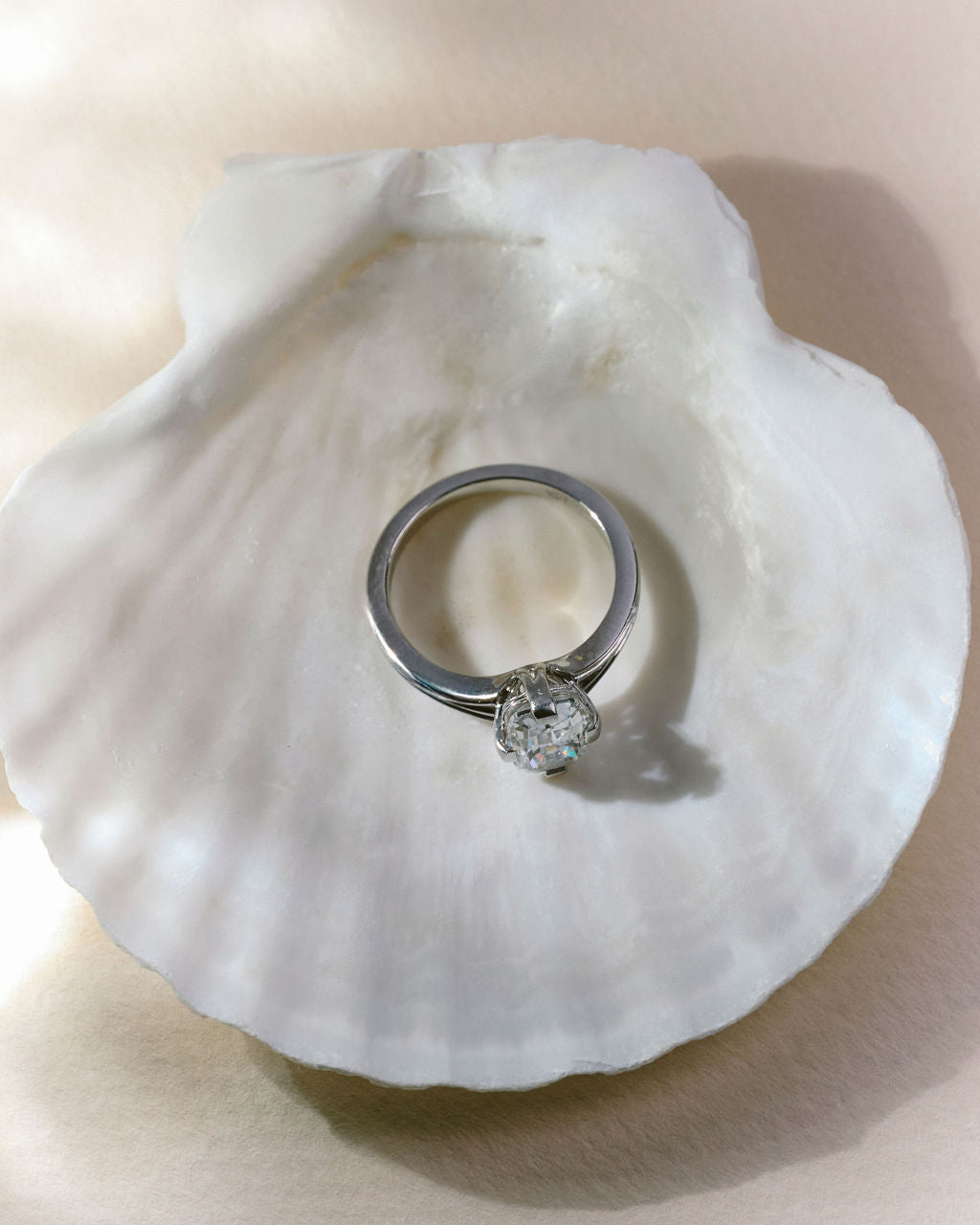 A silver solitaire ring with a cushion diamond resting on a white clam shell. (view 10)