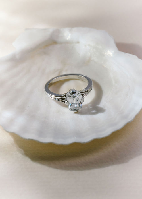 A silver ring with a solitaire gemstone resting on a white clam shell against a warm beige background. (view 4)