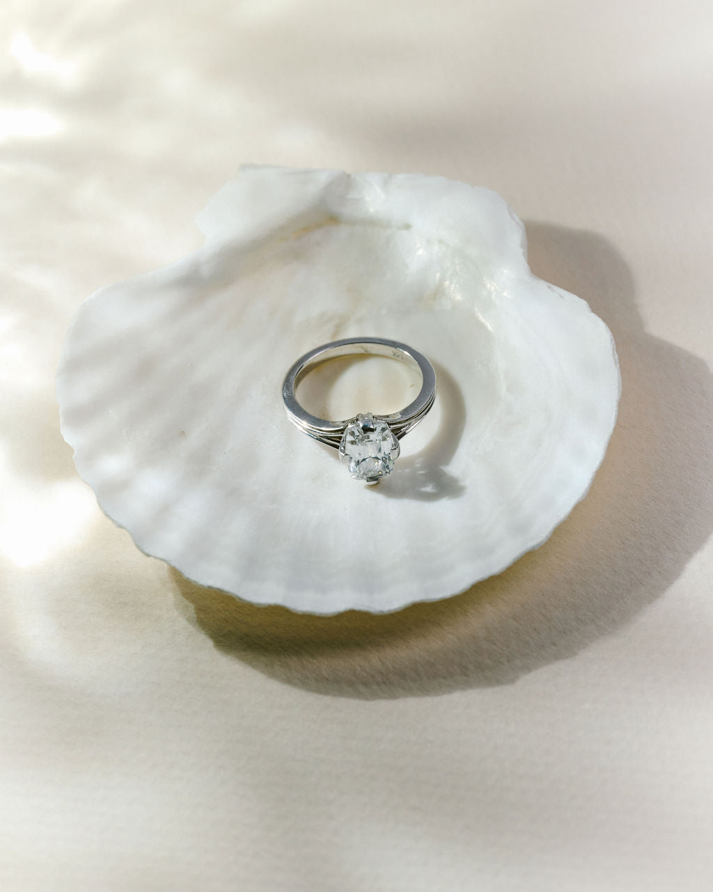 A silver solitaire ring with a cushion gemstone resting on a white clam shell, set against a warm beige fabric background with soft shadows. (view 8)