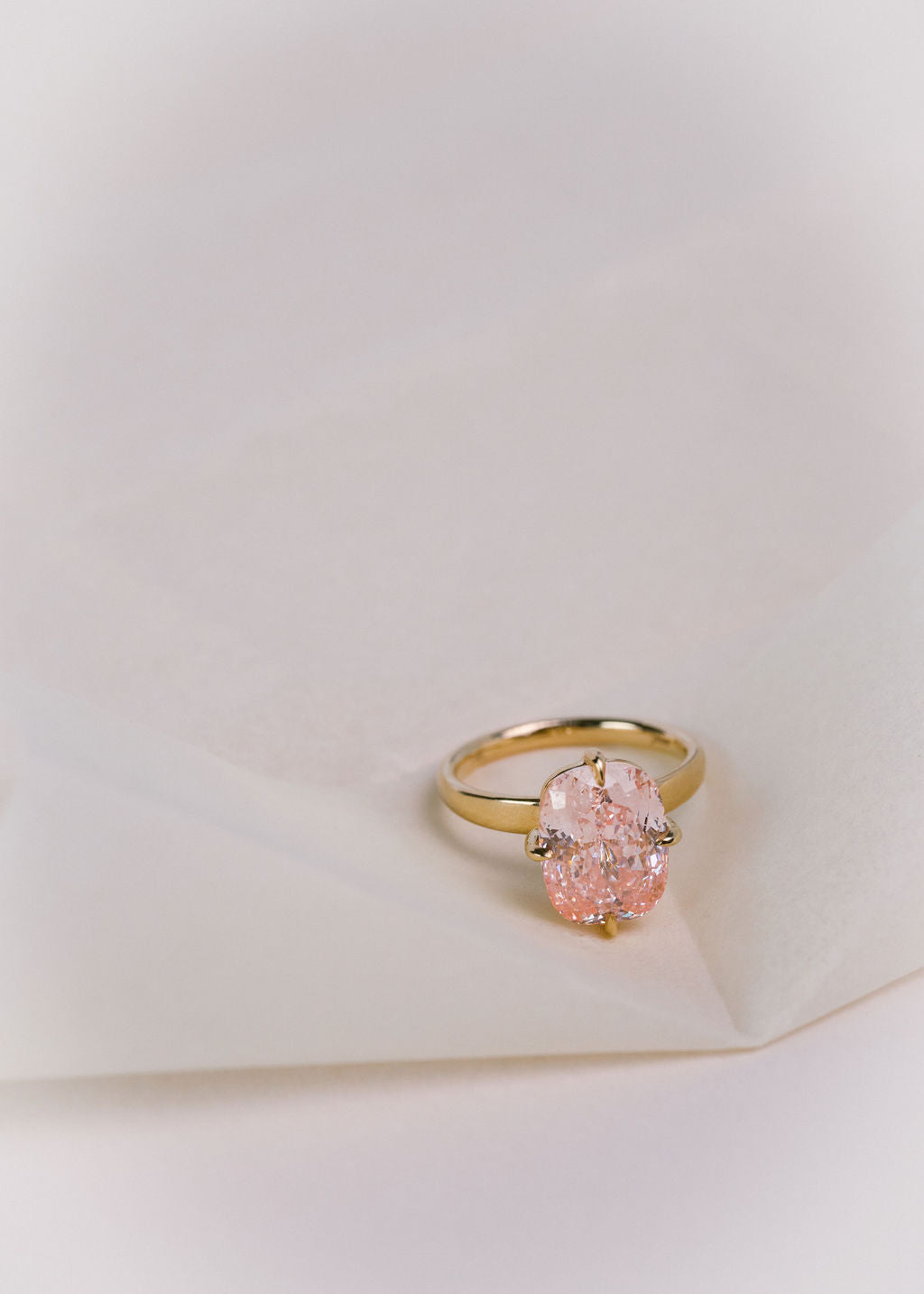 A gold ring with a large, pink cushion diamond, resting on a soft, textured white surface. (view 10)