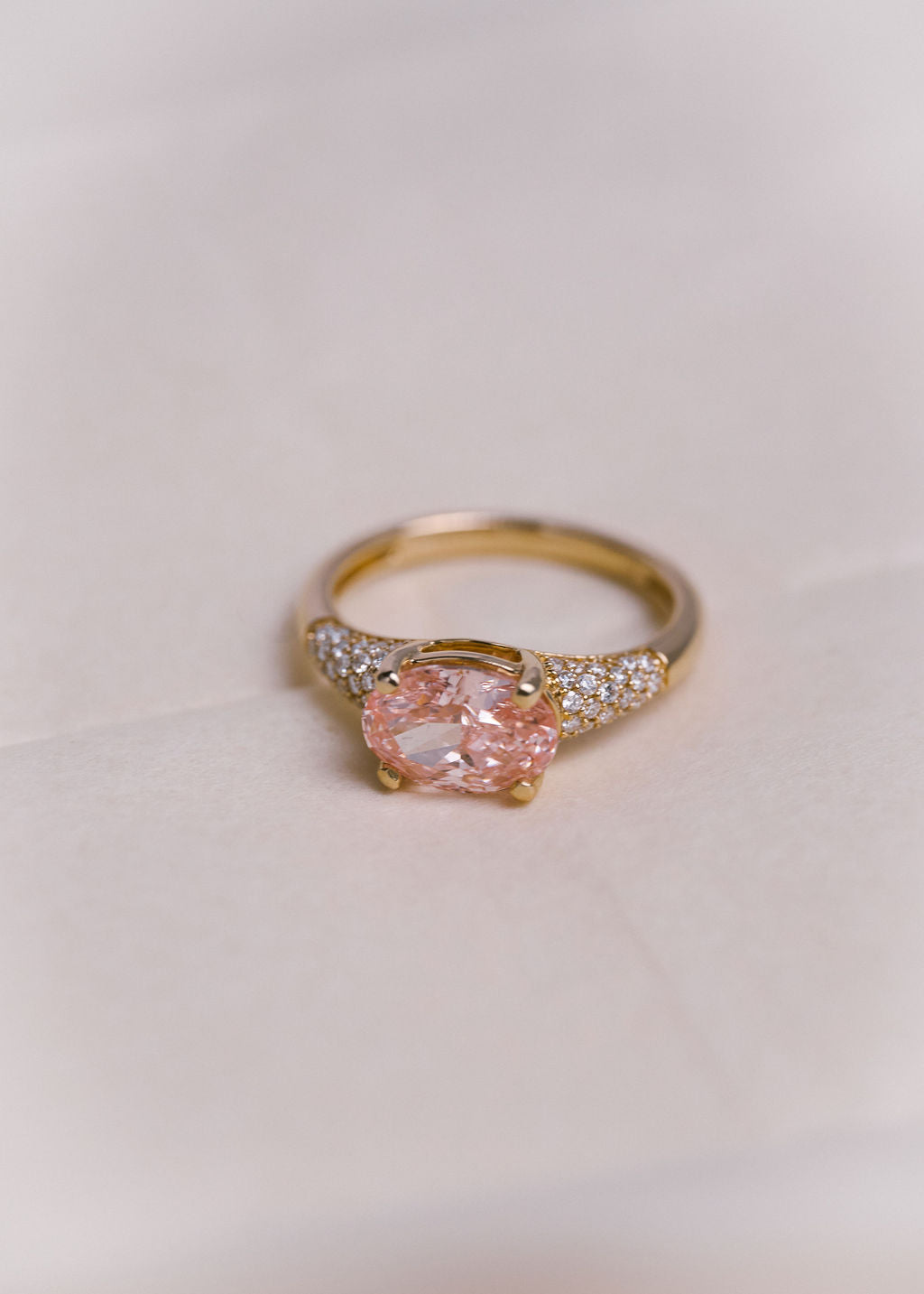A gold ring featuring a large oval pink gemstone in a prong setting, with pavé diamond accents on the shoulders, photographed on a soft off-white background.