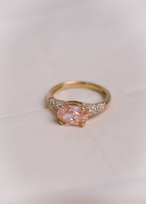 A gold ring featuring a large oval pink gemstone in a prong setting, with pavé diamond accents on the shoulders, photographed on a soft off-white background.